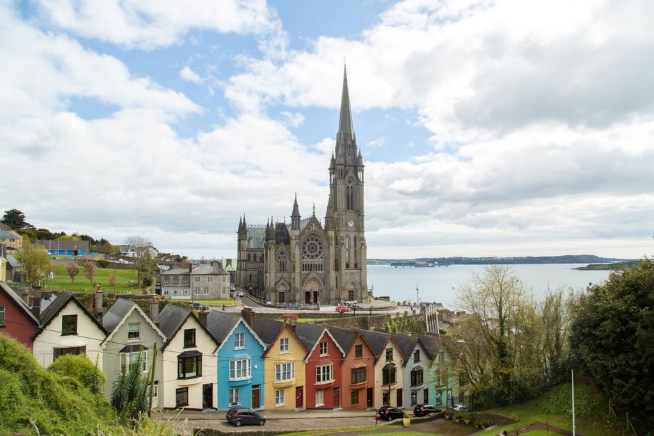 A panoramic view of Cork city centre showing rows of colorful terraced houses in pastel shades of white, blue, yellow, red, and green positioned along a hillside, with a historic Gothic-style church f