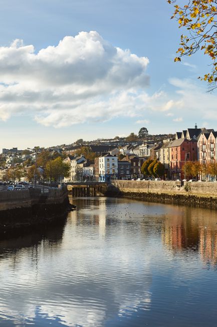 A panoramic view of Cork city centre showing rows of colorful terraced houses in pastel shades of white, blue, yellow, red, and green positioned along a hillside, with a historic Gothic-style church f