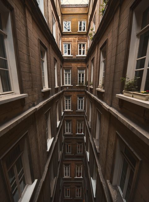 A narrow, enclosed courtyard between multi-storey stone and brick residential buildings with multiple white-framed windows on each floor. The courtyard is vertically deep, with several floors visible,